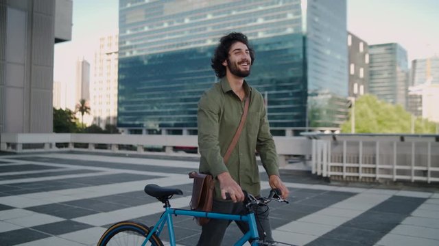 Side view of a smiling handsome young businessman walking outdoors near office buildings with his blue bicycle