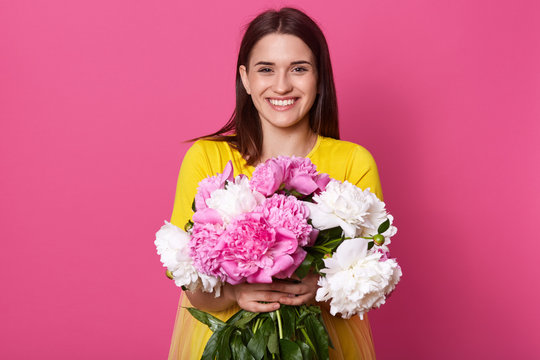 Indoor shot of nice young woman holding beautiful fresh blossoming bouquet of white and pink peony flowers, lady wearing yellow dress, posing isolated over rose background, looking smiling at camera.