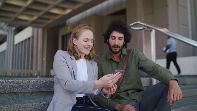 A attractive well dressed woman in business clothing shows a handsome man her smart phone sitting together on office building staircases
