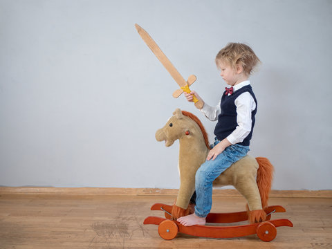 Funny child in white shirt with bow-tie, on toy rocking horse with wooden sword. boy dreams of battles, victories and adventures. concept of education of spirit, education of morale, patriotism