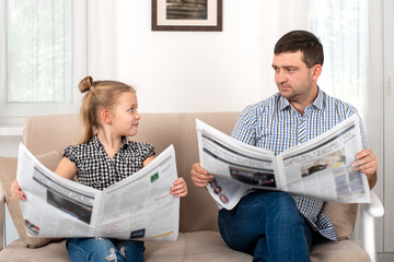 Young girl with  her dad sitting on the sofa at home and reading the newspaper together same way.  Daughter imitates her father.