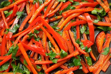 Slices of carrot and red sweet pepper are sauteing in a black frying pan