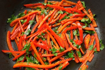 Slices of carrot and red sweet pepper are sauteing in a black frying pan