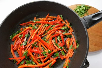 Slices of carrot and red sweet pepper are sauteing in a black frying pan
