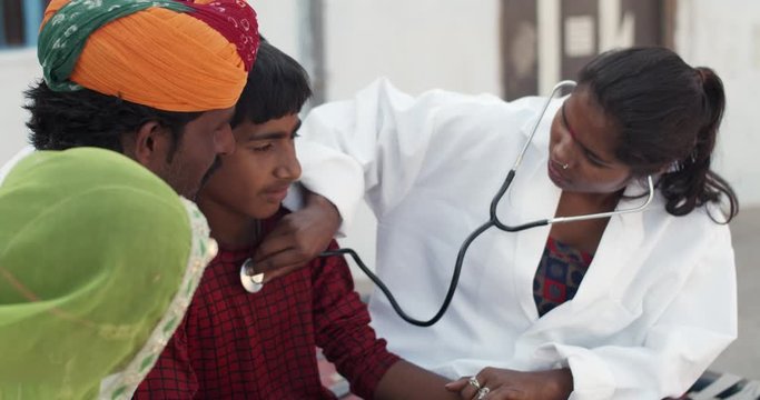 Young medical inter female student using a stereoscope to check the heartbeat of teenage male child in rural Rajasthan, India with his father and mother concerned about his well being and health