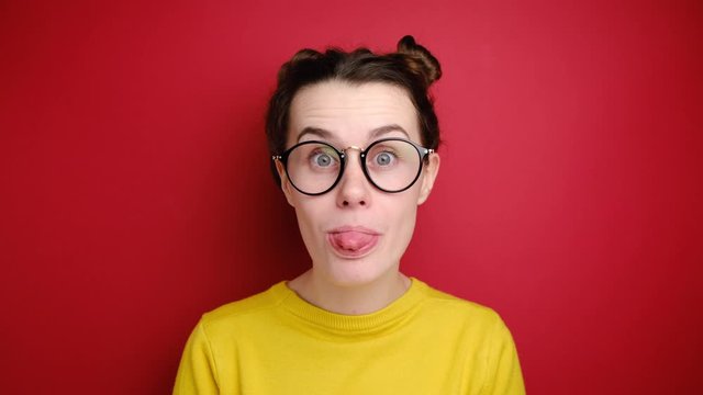 Comic young woman in glasses makes funny face, crosses eyes, plays fool, doesnt want to be responsible, wears yellow sweater, stands over red background. Facial expressions, joy, amusement concept