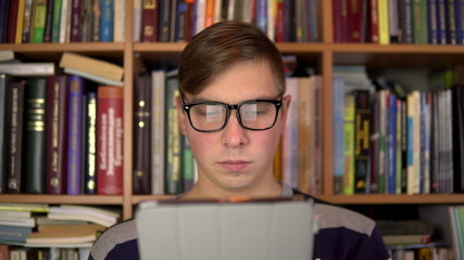 A young man is reading a book in a tablet. A man with glasses carefully looks at the tablet. In the background are books on bookshelves. Book library.