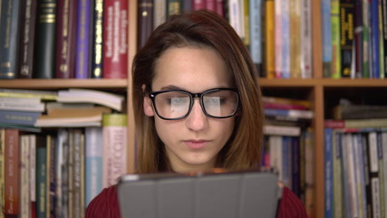 A young woman is reading a book in a tablet. A woman with glasses carefully looks at the tablet. In the background are books on bookshelves. Book library.