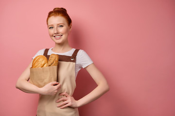 Redhead girl with gathered hair, in an apron, stands on a pink background with a paper bag with bread, holding hand on the waist