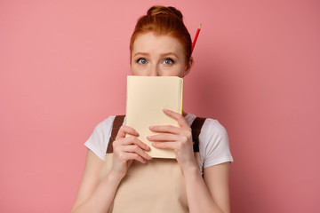 A red-haired girl in an apron with collected hair stands on a pink background, covering her mouth with a notebook and wide-eyed