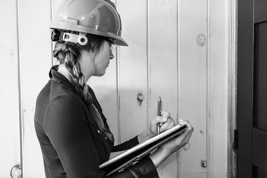 Worker Woman Wears A Grey Hardhat At Work. Female Construction Inspector Taking Notes On Her Clipboard During A Home Inspection, Black And White.