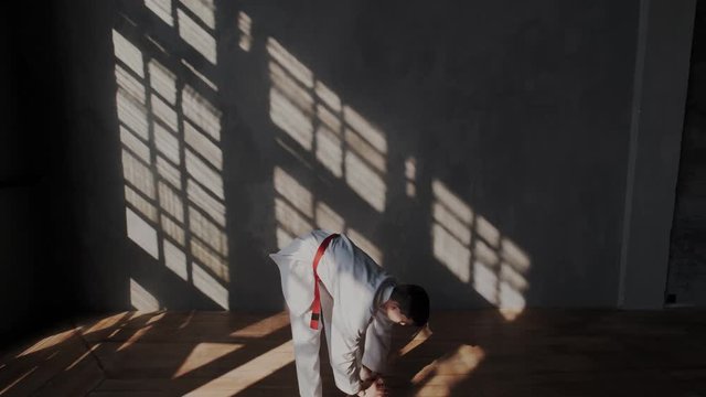 A young brave and strong sportsman is preparing for a taekwondo duel. White kimono with a red belt, barefoot, sunlight from the window, training room. Camera zoom out from boy.