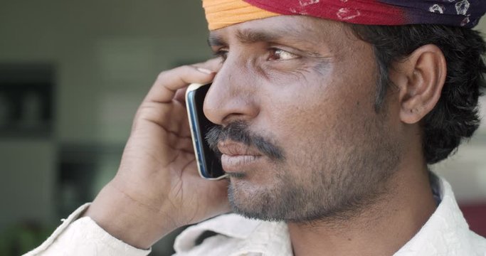 Close-up of young Indian man wearing a traditional colourful Rajasthani turban as she talks and communicates on his smart phone mobile device, positive and cheerful look straight at camera