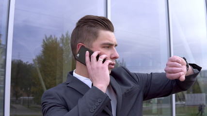 A young businessman in a suit speaks on the phone. Serious man stands in front of a mirror business center