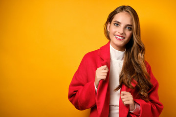 A beautiful girl with clean skin and red lipstick stands on a yellow background and corrects the lacquers of a red coat