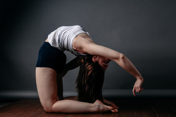 Beautiful woman standing in camel pose on a wooden floor doing Ustrasana exercises, practicing yoga, a sports girl in black sports shorts and a top, exercising home in yoga studio with gray background
