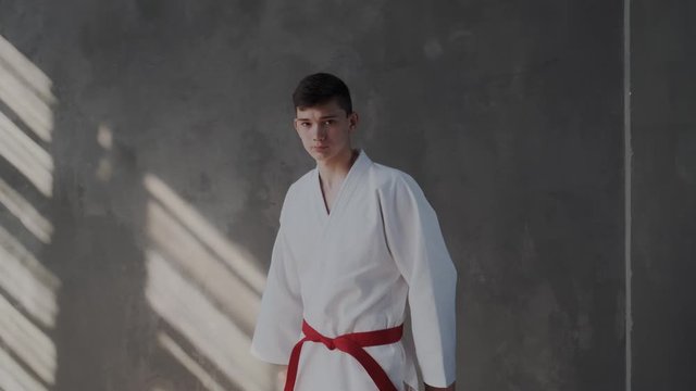 Young fighter belted in a red belt. White kimono, collected look into the camera. The guy is tying a knot and will raise his head to the camera while she is approaching and zoom in to close up.