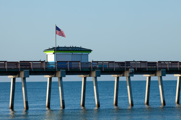 A section of the Venice (FL) municipal fishing pier is shown highlighting the bait shop. An American flag is waving in the breeze and several pigeons are on the roof of the bait shop.