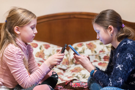 Two girls on the bed play on smartphones. sisters, are lying in bed and looking at a smartphone against the background of the sun from the window. Persian girls on the bed with a phone
