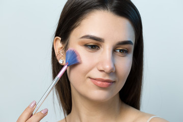 Close-up of a young girl in a light top on a white background making a facial make-up. A pretty woman holds a cosmetic brush near her face and smiles.