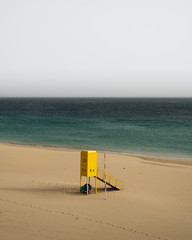 Very calm shot of beach with textured sand hills, blue water and yellow rescue post