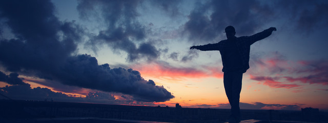 Silhouette of a young active man with outspread hands standing on top of the roof in the city at sunset. Wide screen