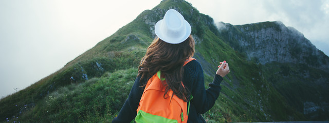 Young traveling woman with backpack sitting high on the top of the mountain with hat and waving hair