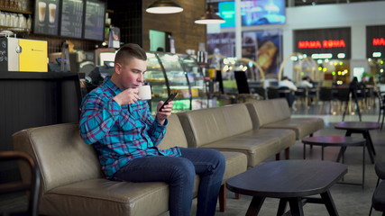A young man is sitting in a cafe with a phone. The man is in correspondence on a smartphone and drinks coffee. Close-up.