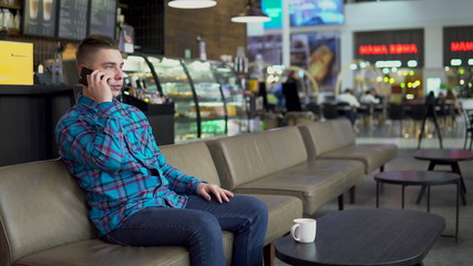 Obraz premium A young man is sitting in a cafe with a phone. The man is in correspondence on a smartphone and drinks coffee. Close-up.