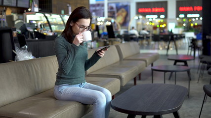 A young woman is sitting in a cafe with a phone. The girl is in correspondence on a smartphone and drinks coffee. Close-up.