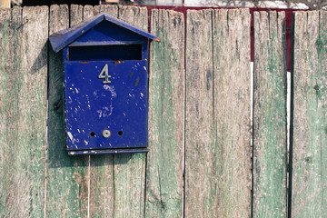 Old gray fence with peeling, green paint and a blue dirty mailbox with number 4. Aged boards with slots close-up