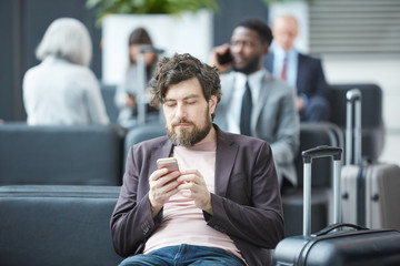Horizontal portrait of young Caucasian man sitting relaxed in departure lounge surfing Internet on his smartphone