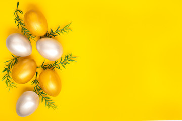 Naklejka na ściany i meble Eggs of silver and gold, green twigs from the left edge of the image on a plain yellow background. Easter holiday card. Copyspace.