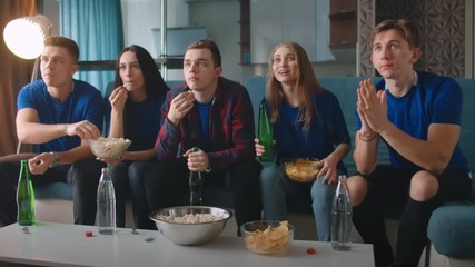 5 people men and women in blue t-shirts are emotionally worried watching TV sitting at home on the couch, with popcorn and drinks, happy to score a goal.