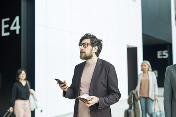 Horizontal medium portrait of pensive young man standing in airport hall holding smartphone, boarding pass and purse looking at information display