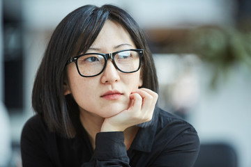 Cloe up head and shoulders portrait shot of attactive young Asian woman wearing stylish eyeglasses resting chin on her hand looking away