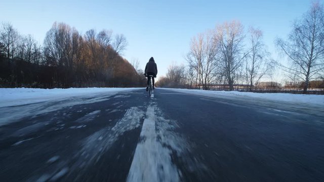 Man swerving with his bicycle on empty snowy road. Tracking shot from behind