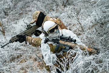 Two men in camouflage white, green uniform with machineguns back to back. Soldiers with muchinegun stood on knelt in the winter grass.