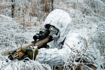 Military man in white camouflage uniform with hood and machinegun in the long winter grass. Soldier...