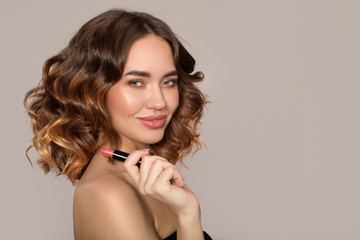 Portrait of a smiling young beautiful fashion woman. Volumetric hairstyle with wavy hair and makeup. Holding lipstick in hand.