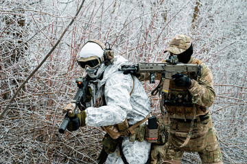 Fototapeta premium Two men in camouflage white, green uniform with machineguns. Soldiers with muchineguns go one after another in the winter forest. Horizontal photo