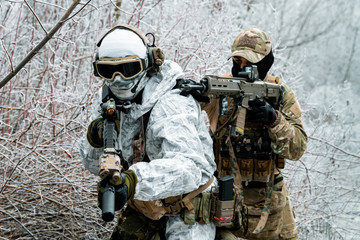 Closeup two men in camouflage white, green uniform with machineguns. Soldiers with muchineguns go one after another in the winter forest.