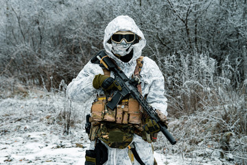 Military man in white camouflage uniform with hood and machinegun. Soldier in the winter forest...