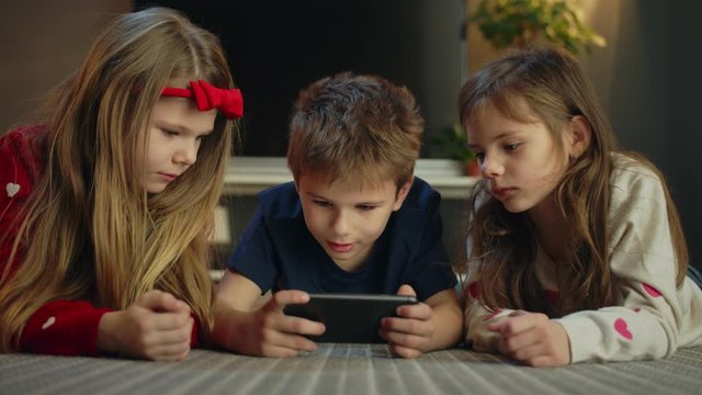 Portrait of Caucasian brother and twin sisters watching video online using smartphone, young family generation staying online together holding mobile device enjoying cartoon laying on floor at home
