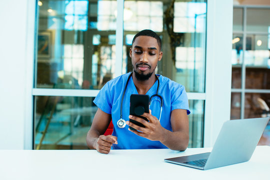 Portrait of a concerned male doctor or nurse wearing blue scrubs uniform and stethoscope sitting at desk with laptop in hospital checking mobile phone