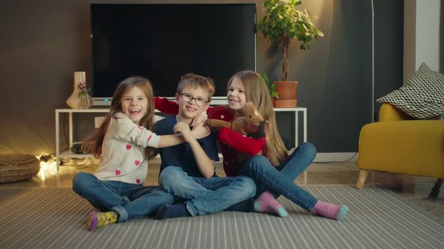 Cheerful Caucasian lovely young kids triplets having fun together in modern living room, attractive little brother and sisters kissing and hugging sitting on floor carpet in casual jeans indoors
