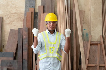 Handsome man wearing protective glasses and yellow helmet,raised fists up with determind eyes,standing in front of wood pile,at factory
