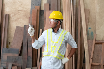 Handsome man wearing protective glasses and yellow helmet,point finger to right side,standing in front of wood pile,at factory