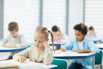 Horizontal portrait of little blond girl with two plaits sitting at school desk resting her cheek on hand learning boring lesson, copy space