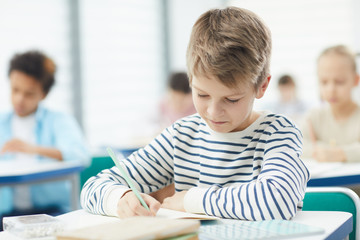 Fototapeta na wymiar Caucasian boy wearing striped sweatshirt sittng at desk in classroom doing lesson task in his notebook, horizontal medium close up portrait, copy space